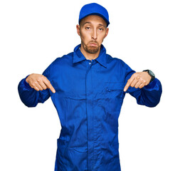 Bald man with beard wearing builder jumpsuit uniform pointing down looking sad and upset, indicating direction with fingers, unhappy and depressed.