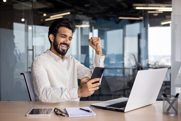 Muslim young man sitting in the office at the desk, looking at the phone and showing a victory gesture with his hand - obrazy, fototapety, plakaty