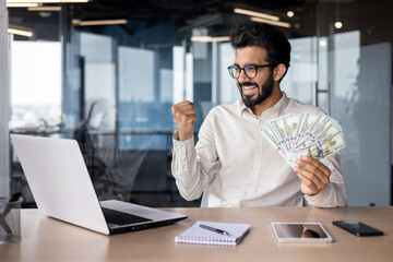 Young indian man working in office at laptop, holding money cash, happy with success, showing victory gesture with hand