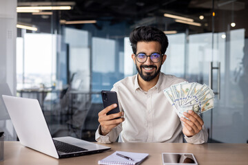 Portrait of a young Indian man sitting in the office at the table, holding cash money and a phone in his hands, smiling and looking at the camera - obrazy, fototapety, plakaty