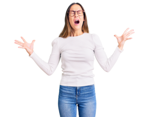 Beautiful brunette young woman wearing casual white sweater and glasses celebrating mad and crazy for success with arms raised and closed eyes screaming excited. winner concept