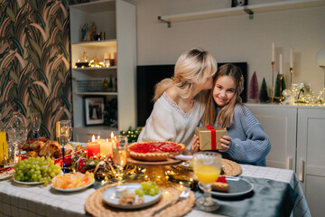 Portrait of loving mother hugging smiling little daughter after giving birthday present to adorable daughter sitting at dinner feast table during family party. Concept of home holiday atmosphere.