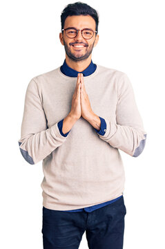 Young handsome hispanic man wearing elegant clothes and glasses praying with hands together asking for forgiveness smiling confident.
