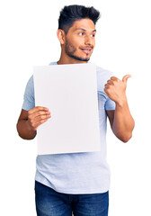 Handsome latin american young man holding cardboard banner with blank space pointing thumb up to the side smiling happy with open mouth