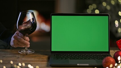 Glass and bottle of red wine with laptop computer on table in Christmas decorated home. Man tasting buying wine online. Green screen for web shop ads and vinery holiday offer. - obrazy, fototapety, plakaty