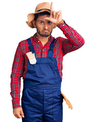 Handsome latin american young man weaing handyman uniform making fun of people with fingers on forehead doing loser gesture mocking and insulting.