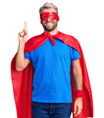 Naklejka premium Young blond man wearing super hero custome showing and pointing up with finger number one while smiling confident and happy.