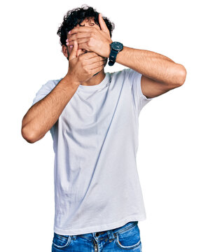Hispanic young man with beard wearing casual white t shirt covering eyes and mouth with hands, surprised and shocked. hiding emotion