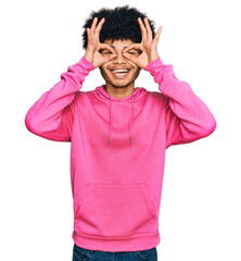 Young african american man with afro hair wearing casual pink sweatshirt doing ok gesture like binoculars sticking tongue out, eyes looking through fingers. crazy expression.