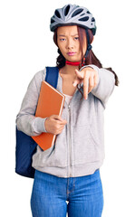 Young beautiful chinese girl wearing student backpack and bike helmet holding book pointing with finger to the camera and to you, confident gesture looking serious