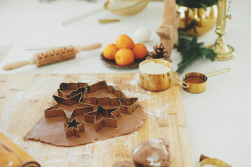 Gingerbread dough with festive golden metal cutters on wooden board with flour, cooking spices, festive decorations in modern white kitchen. Making christmas gingerbread cookies