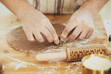 Hands cutting gingerbread dough with festive golden metal cutters on wooden board with flour,...