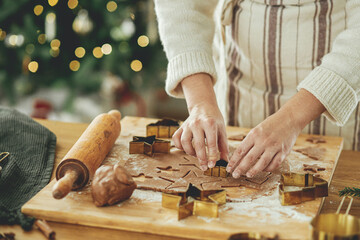 Hands cutting gingerbread dough with festive golden cutters on rustic table with holiday...