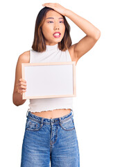 Young beautiful chinese girl holding empty white chalkboard stressed and frustrated with hand on head, surprised and angry face