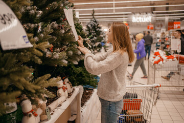 Cute young woman choosing artificial Christmas tree in Xmas store. Christmas concept store, buying...