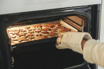 Woman in oven mitts putting tray with fresh gingerbread cookies in christmas festive shapes in...
