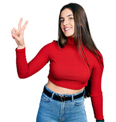 Young brunette teenager wearing red turtleneck sweater smiling looking to the camera showing fingers doing victory sign. number two.