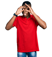 Young african american man wearing casual red t shirt doing ok gesture like binoculars sticking tongue out, eyes looking through fingers. crazy expression.