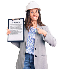 Young beautiful brunette woman wearing architect hardhat holding contract smiling happy pointing with hand and finger