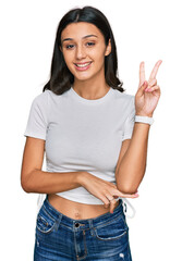 Young hispanic girl wearing casual white t shirt smiling with happy face winking at the camera doing victory sign with fingers. number two.