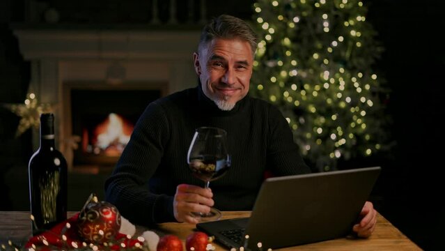 Happy smiling middle aged man at home tasting wine in cosy room with fireplace in winter. Laptop computer, Christmas decor, gift boxes. Concept for web shop ads and vinery holiday offer.