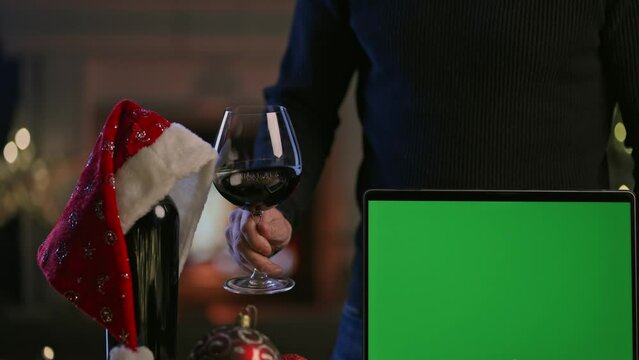 Glass and bottle of red wine with laptop computer on table in Christmas decorated home. Man tasting buying wine. Green screen for web shop ads and vinery holiday offer.
