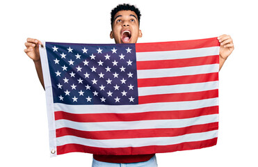 Young african american man holding united states flag angry and mad screaming frustrated and furious, shouting with anger looking up.