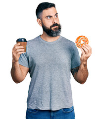 Hispanic man with beard eating doughnut and drinking coffee smiling looking to the side and staring...