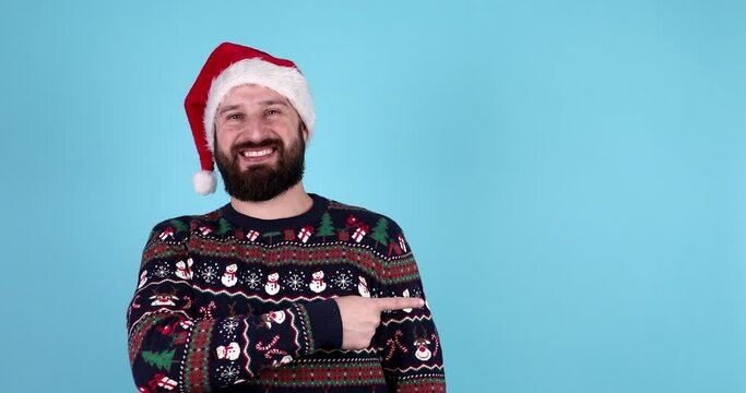 Young bearded male in Santa hat and warm christmas sweater is pointing up at something, winking and smiling while posing against blue background. Happy New Year, advertising. Close up, copy space. 