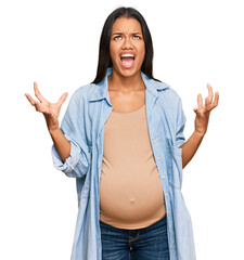 Beautiful hispanic woman expecting a baby showing pregnant belly crazy and mad shouting and yelling...