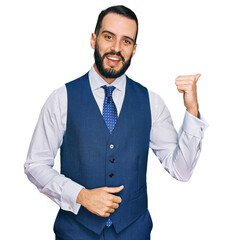 Young man with beard wearing business vest smiling with happy face looking and pointing to the side with thumb up.