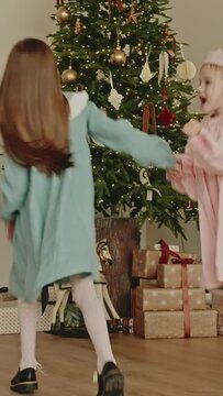 Two happy little sister girls wearing a dresses, dancing in a living room decorated with lights, glowing with xmas tree garlands on Christmas Eve. Happy children celebrate New Year. Vertical video.