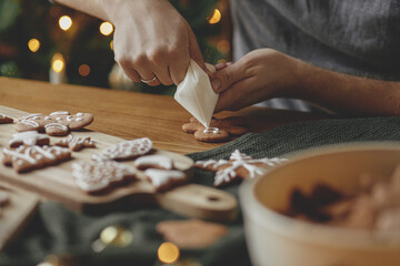Decorating gingerbread cookies with icing on rustic wooden table at christmas tree golden lights....