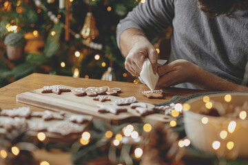 Hands decorating gingerbread cookies with icing on rustic wooden table at christmas tree golden...