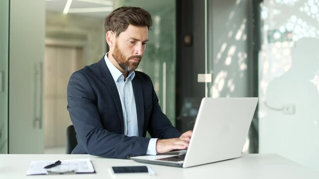 Frustrated businessman in a formal suit having problems with software or service on laptop computer sitting in business office. Worried disappointed worker complains about a bad internet connection
