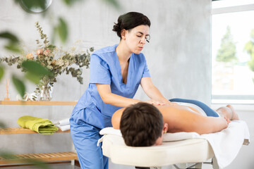 Adult female masseuse makes back massage to young guy client in salon