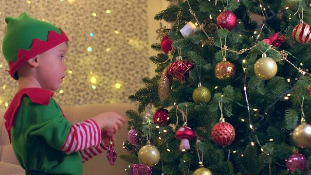 little boy child of 4 years old in green elf costume hangs decorations on Christmas tree. baby elf preparing Christmas and new year celebration waiting for winter holidays with family concept