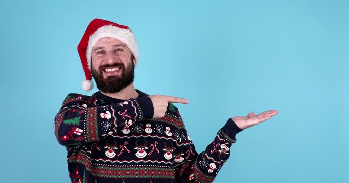 Young attractive smiling bearded man in santa hat posing isolated on blue background. People sincere emotions lifestyle concept. Mock up copy space. Pointing index finger aside.