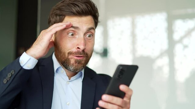 Excited happy businessman in formal suit received great news on smartphone sitting at workplace in business office. A joyful entrepreneur reads pleasant message on phone, celebrates success. Close up