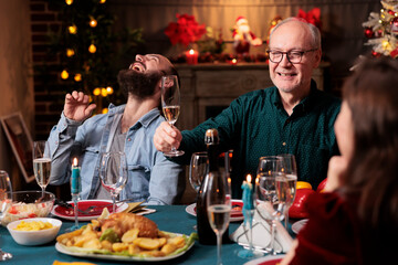 Old adult giving toast with raised glass of champagne at christmas eve celebration to enjoy winter holiday with friends and family. Grandpa making speech around the table, sparkling wine. - obrazy, fototapety, plakaty