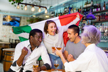 Cheerful international sport fans waving Italian flag and drinking beer in the sport bar - obrazy, fototapety, plakaty