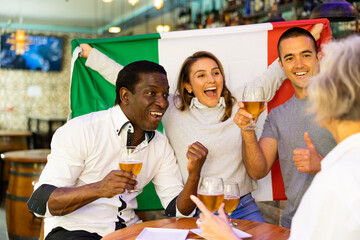 Excited diverse soccer supporters with flag of Italy celebrating victory with pint of beer in the...