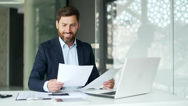 Excited financier in a formal suit is satisfied with results of a financial report he is reviewing sitting at workplace in a business office. Entrepreneur businessman is happy with positive indicators