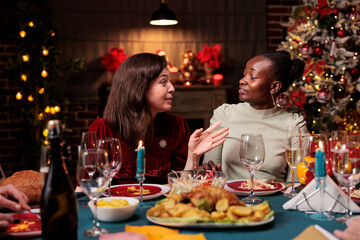 Diverse women celebrating christmas with dinner, enjoying seasonal winter event at home. People having fun with december celebration at table surrounded by festive ornaments and xmas tree.