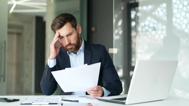 Confused puzzled businessman in a formal suit having difficulty with paper work sitting at workplace in business office. Frustrated financier reviewing documents, unhappy with bad financial results