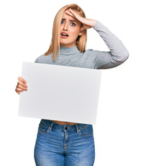 Beautiful caucasian woman holding blank empty banner stressed and frustrated with hand on head, surprised and angry face