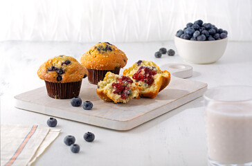  Blueberry muffins on a white cutting board with a glass of milk in the foreground and a bowl of fresh blueberries in the background; copy space