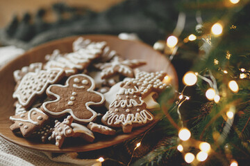 Merry Christmas! Gingerbread cookies with icing in plate on wooden rustic table with fir branches, festive decorations and christmas golden lights. Atmospheric Christmas holidays, family time