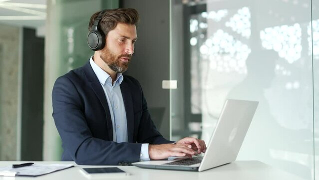 Busy businessman in wireless headphones typing on laptop computer while sitting at workplace at desk in modern office. Handsome entrepreneur in formal suit works on a project while listening to music