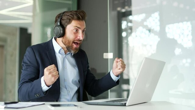 Excited businessman in wireless headphones watching sports match or competition using laptop while sitting at workplace in office Happy man in formal suit cheering for favorite team with emotional ges